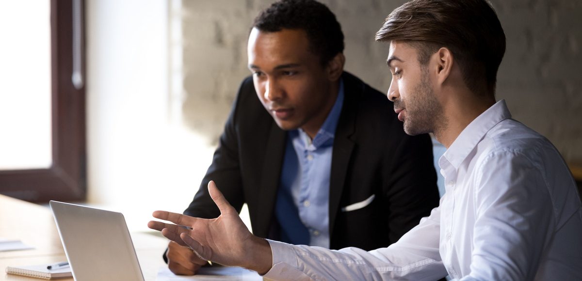 african intern receiving instructions from caucasian colleague sitting using laptop